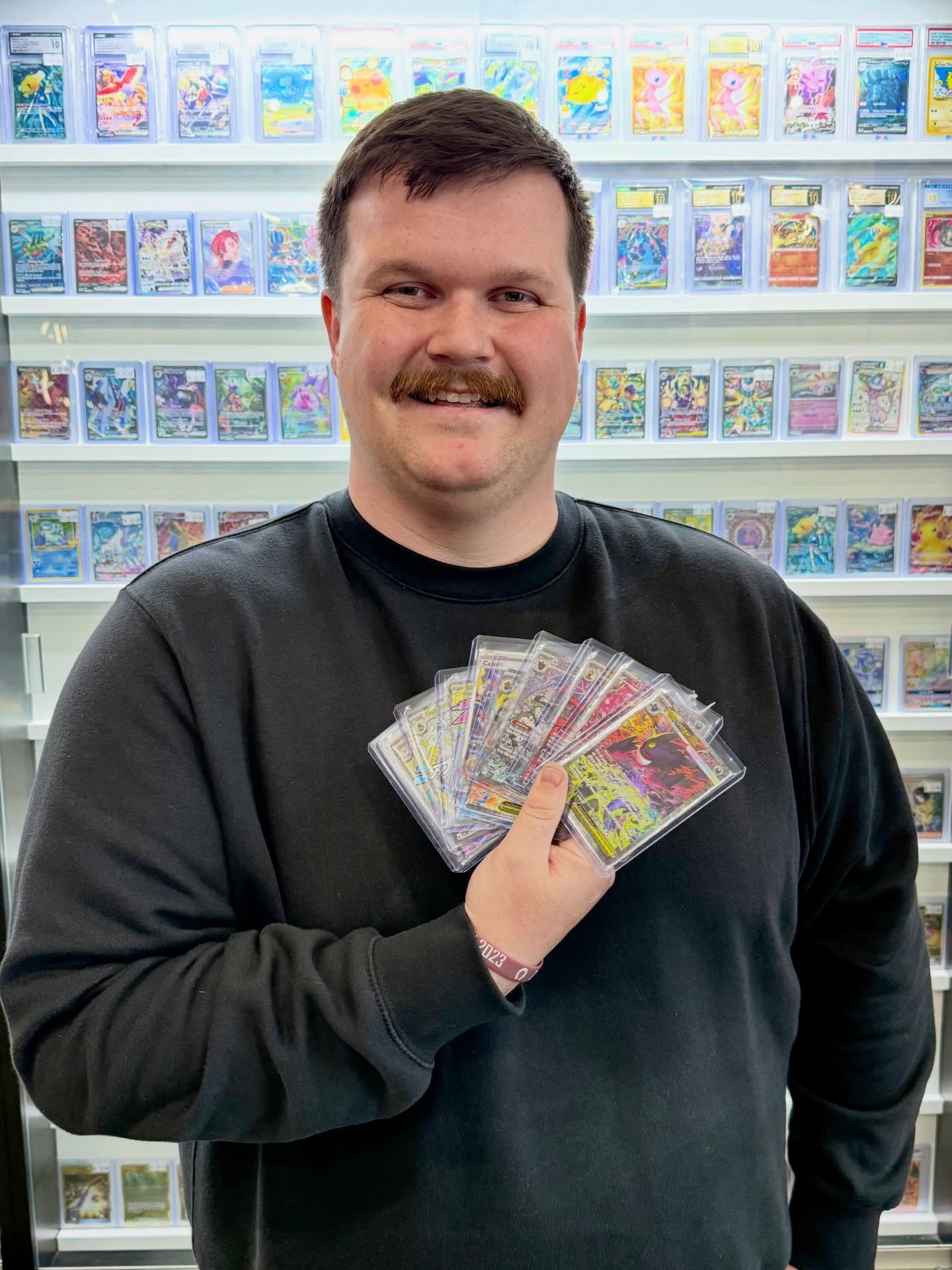 Smiling man in a black sweatshirt holding a fan of trading cards in a card shop.
