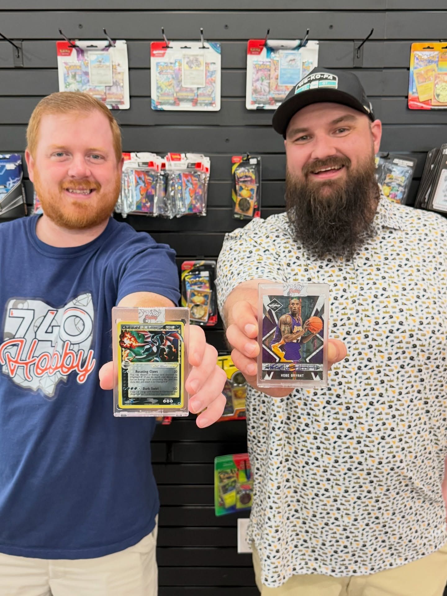 Two men in a store display trading cards to the camera, standing in front of a wall of hanging products.