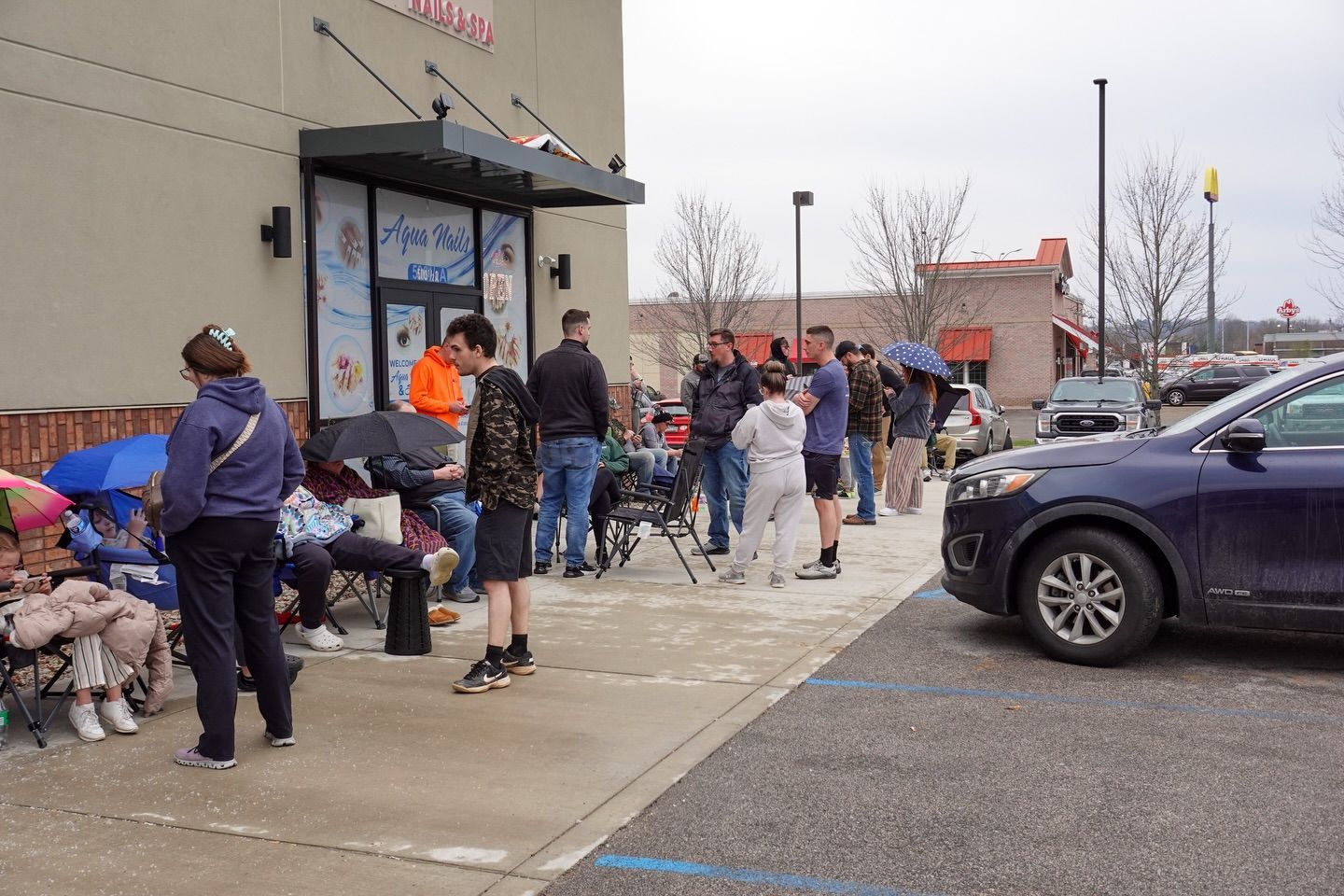 People waiting in line outside a storefront on a chilly day, with a purple SUV parked nearby.