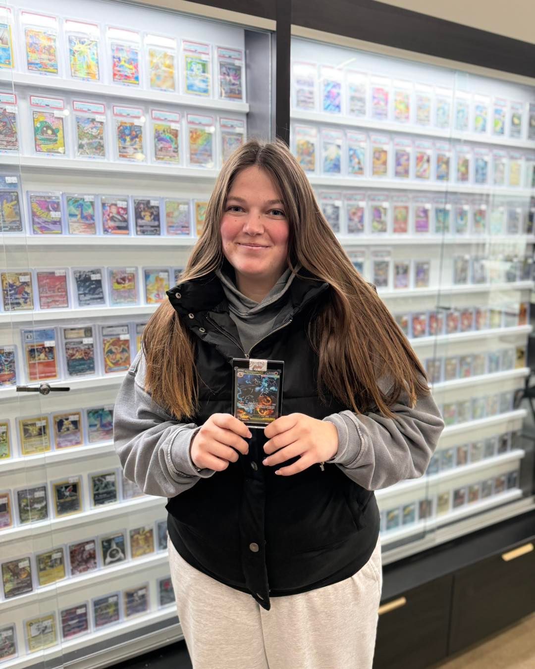 Person holding a trading card in front of a wall of card displays, smiling in a store.