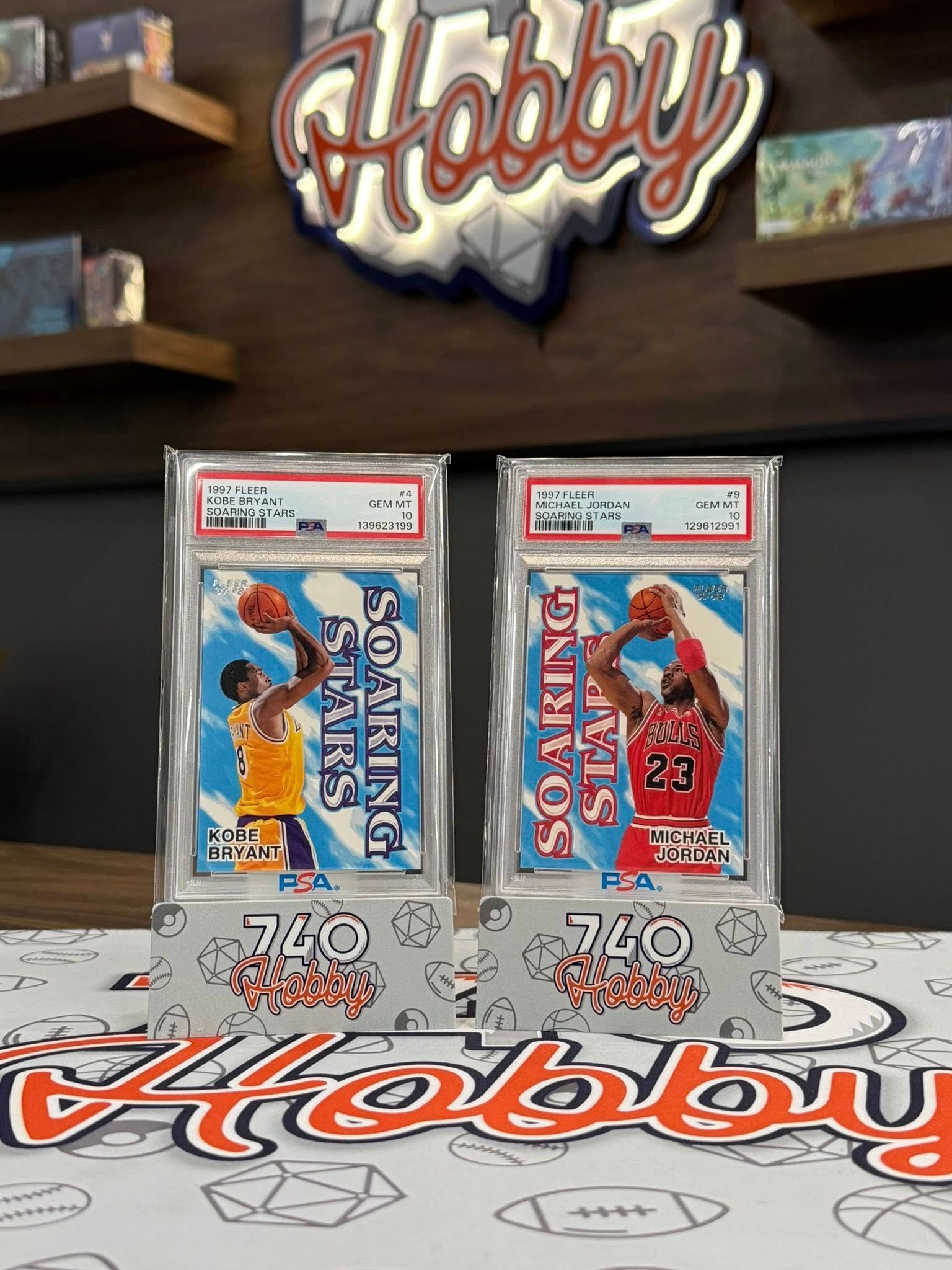 Two graded baseball cards displayed on a Hobby-branded table in front of a shelf backdrop