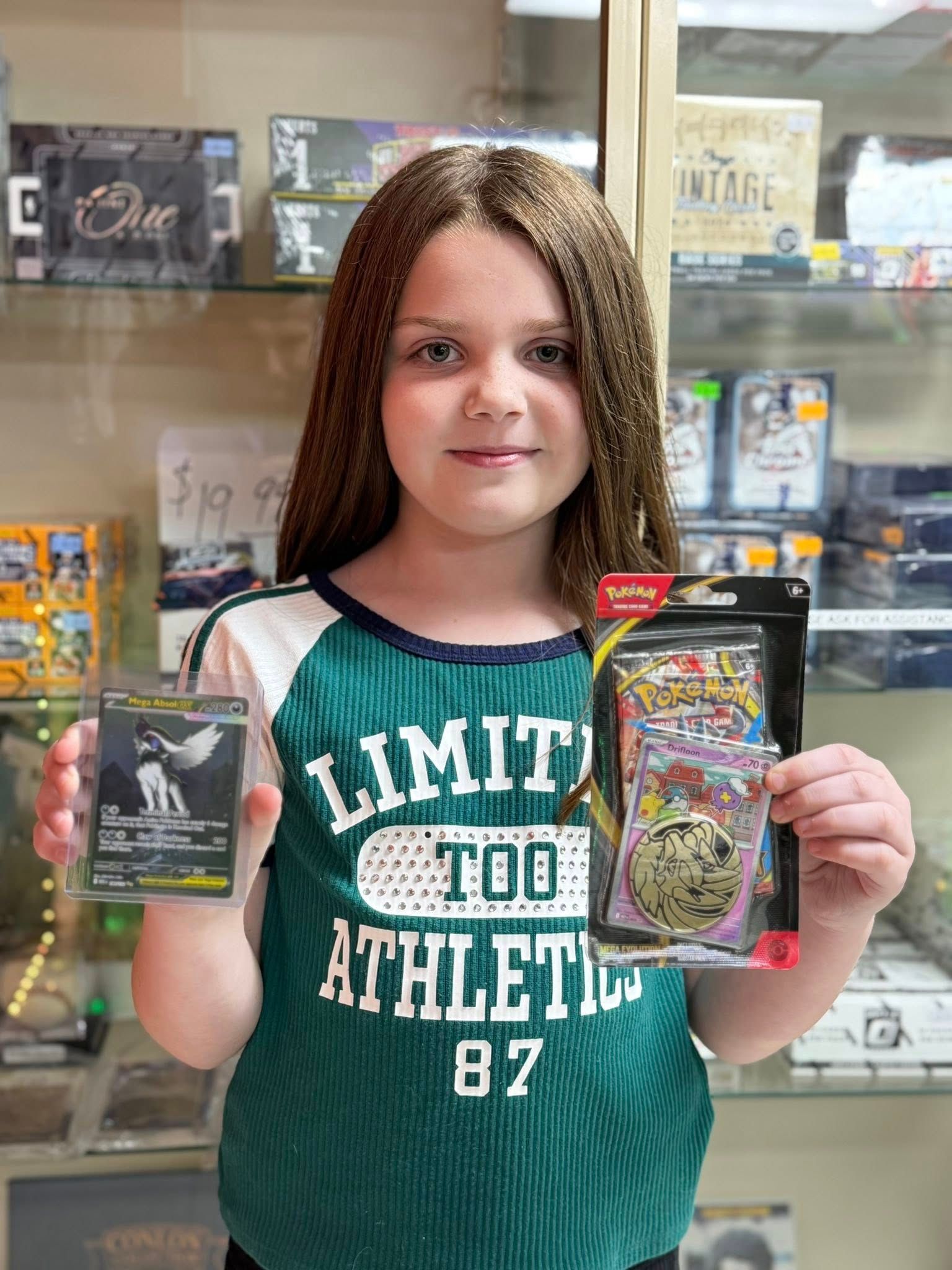 Girl holding two trading card packs in a store aisle, wearing a green “Limited Too Athletic” shirt.