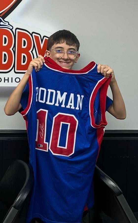 Smiling child holding up a blue and red Rodman #10 jersey in front of a wall sign