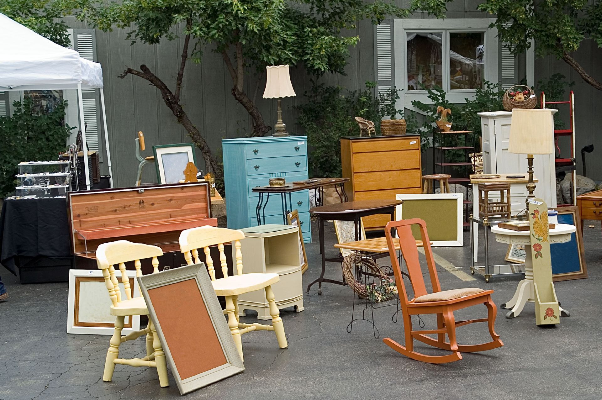 Yard sale with furniture: chairs, chests, lamps, and framed art outside a building.