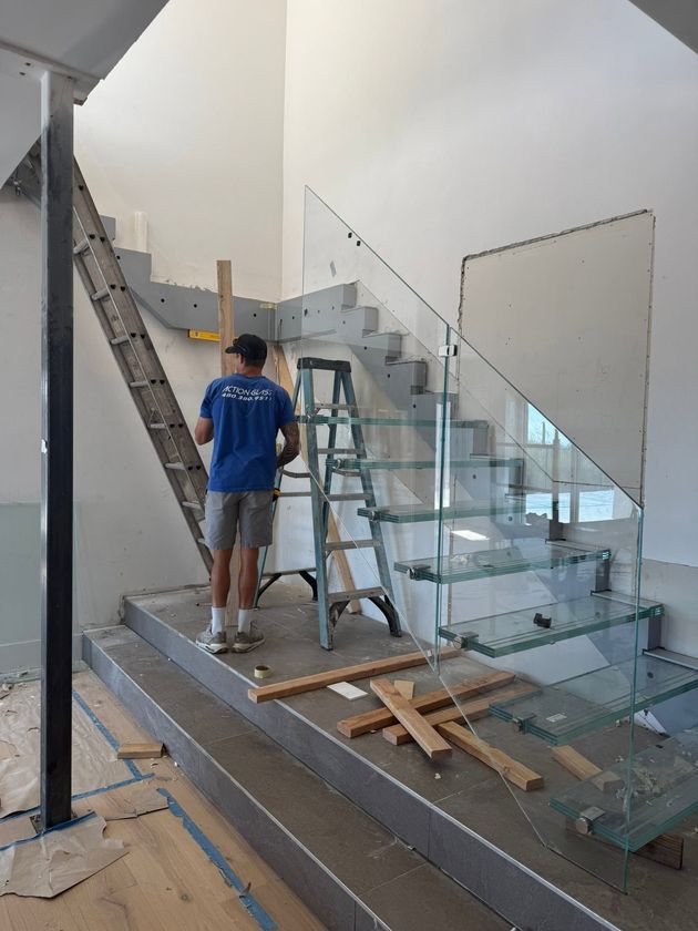 Worker installing a glass staircase in a bright indoor space, with tools and materials on the floor.