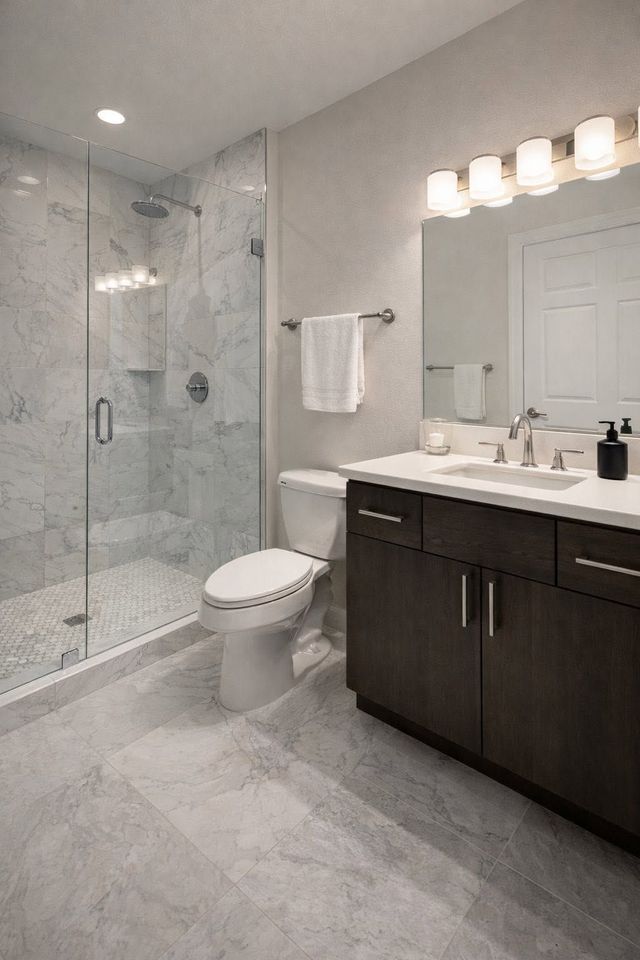 Modern bathroom with glass shower, toilet, dark vanity, and large mirror under bright lights
