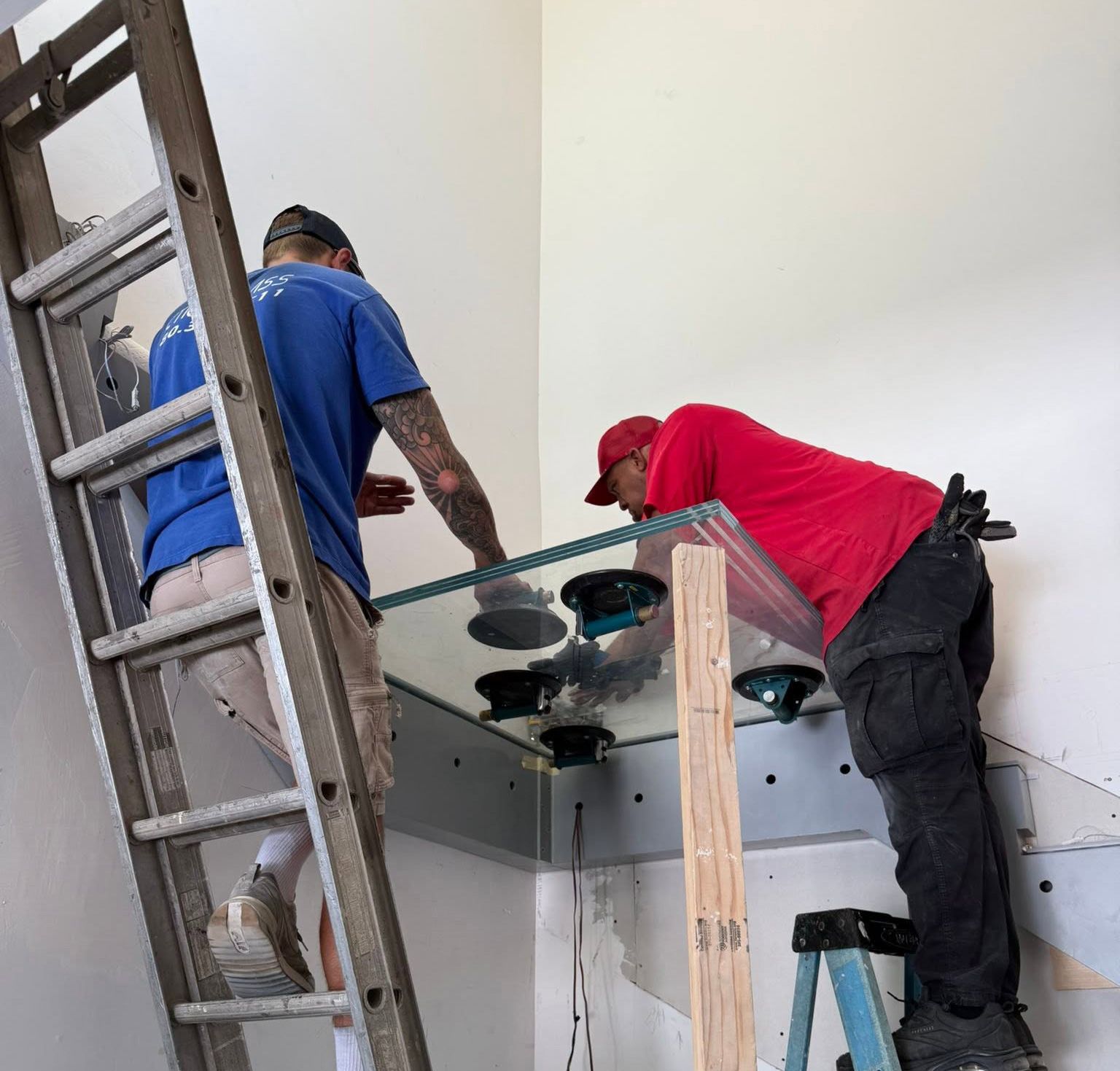 Two workers installing a glass fixture on a wall, one on a ladder and one leaning over the frame.