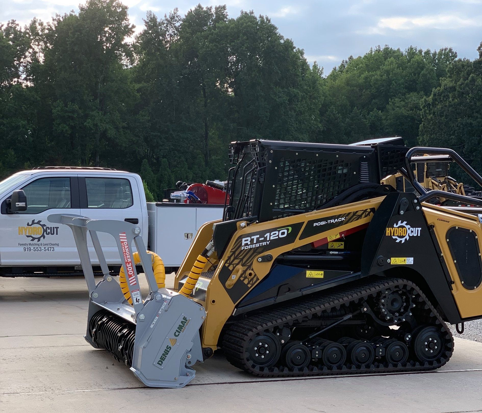Yellow and black track skid steer with a brush cutter attachment parked in front of a white truck.