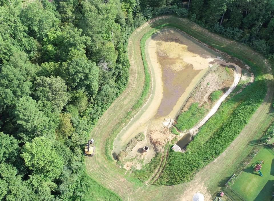 Aerial view of a man-made pond with brown and green banks surrounded by forest and grass.