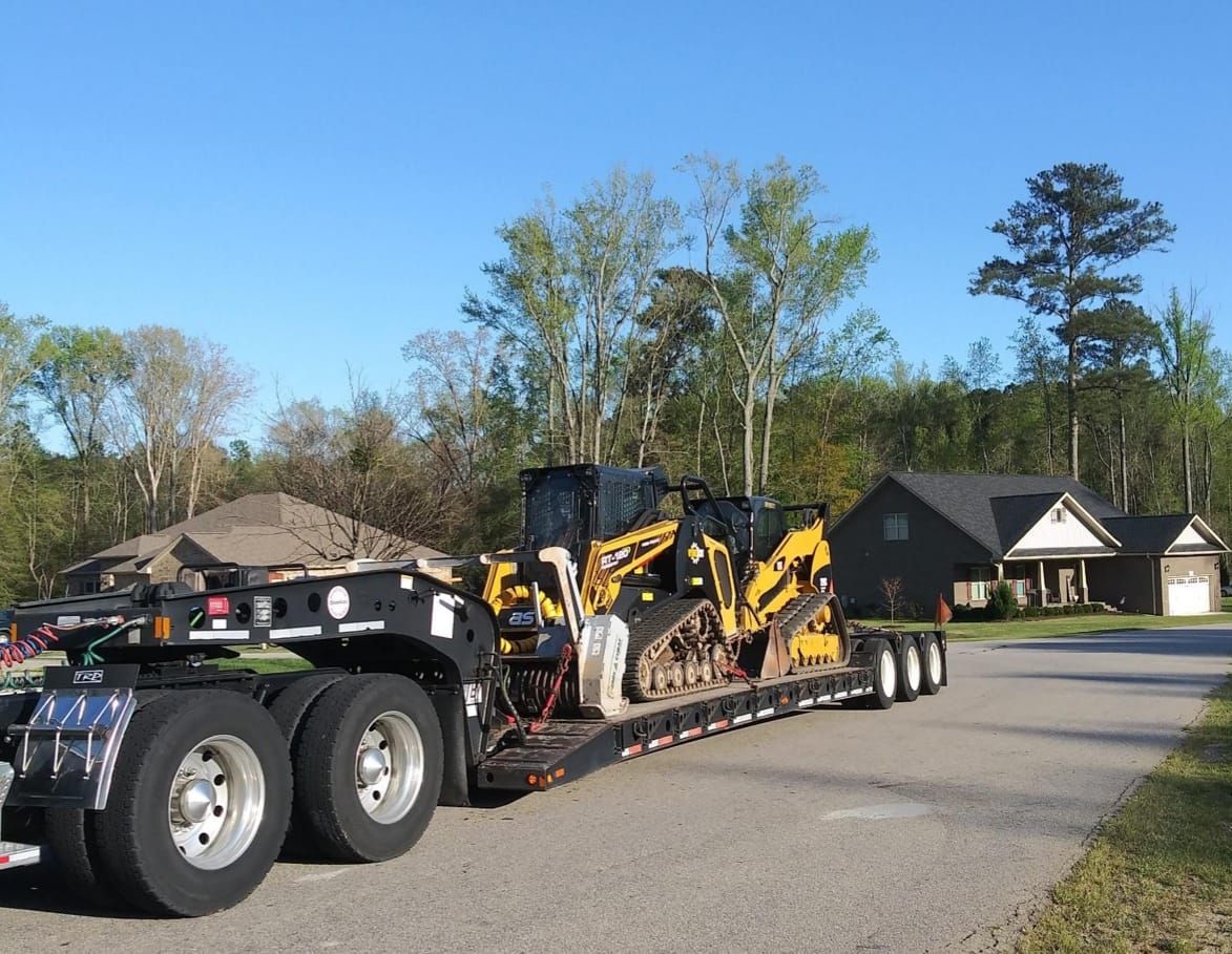 Heavy equipment loaded on a semi-trailer on a residential street, trees and houses in the background.