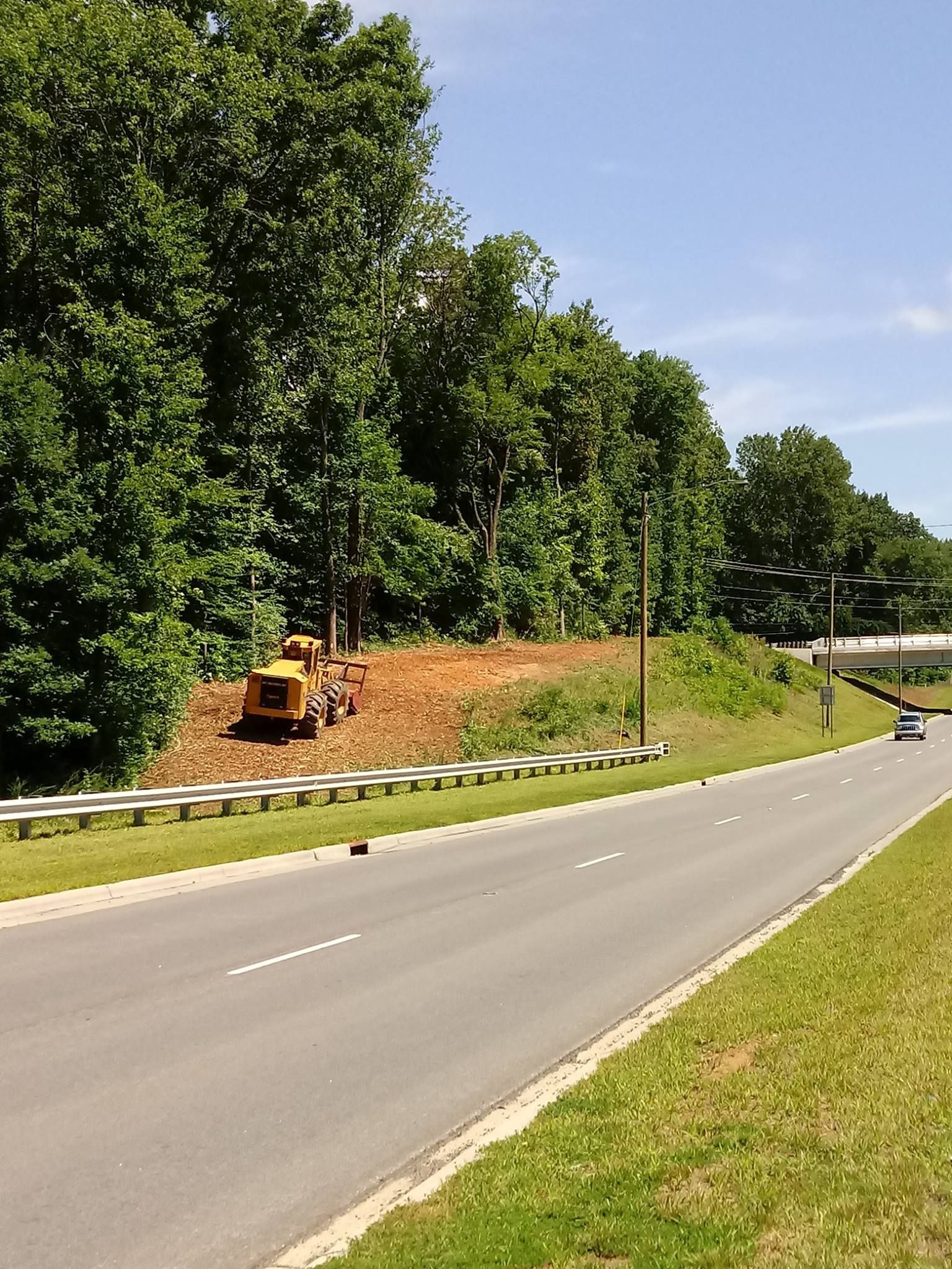 A road with a bulldozer on a cleared hillside next to a forest under a blue sky.