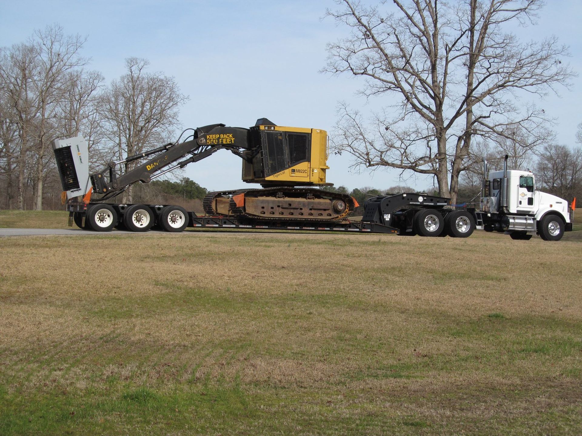 A large yellow excavator on a flatbed trailer is being pulled by a white semi-truck on a grassy field.
