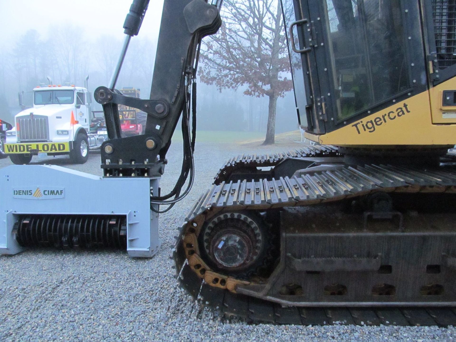Excavator with a grinding attachment next to a semi-truck, outdoors, foggy conditions.