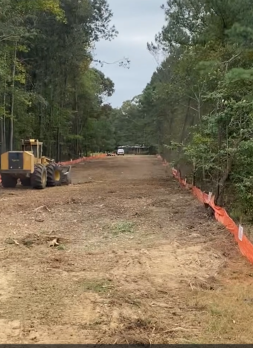 Dirt road cleared through a forest; orange sediment barriers line sides. A yellow construction vehicle sits on the left.