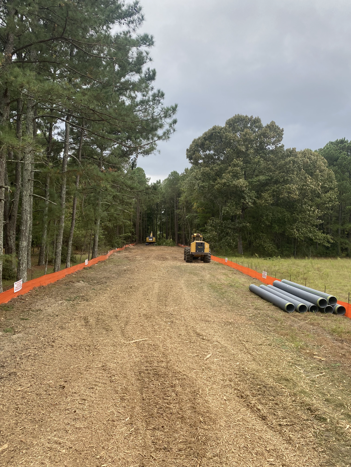 Gravel road through trees with orange safety barriers, construction vehicles, and gray pipes.