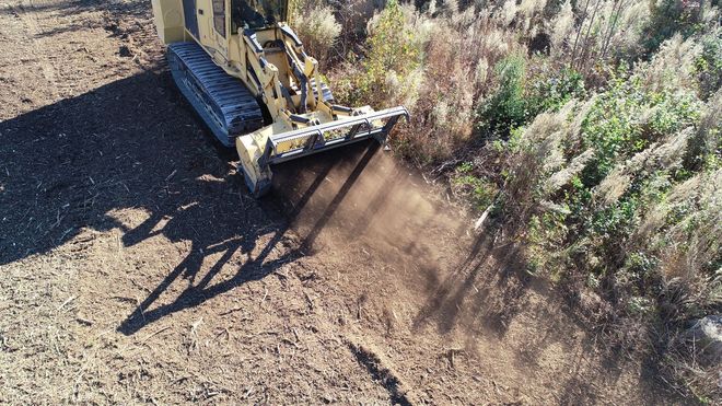 A yellow tracked machine clears brush in a field, kicking up dirt.