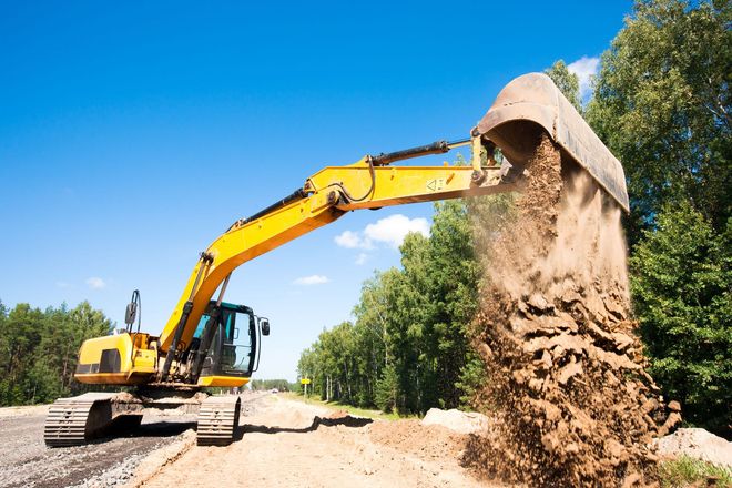 Yellow excavator digging dirt on a roadside under a blue sky.