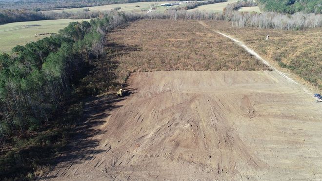 Aerial view of a partially cleared field with brown soil, adjacent to a forest and a dirt road.