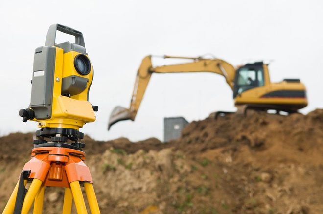A yellow surveying instrument on a tripod in front of an excavator on a construction site.