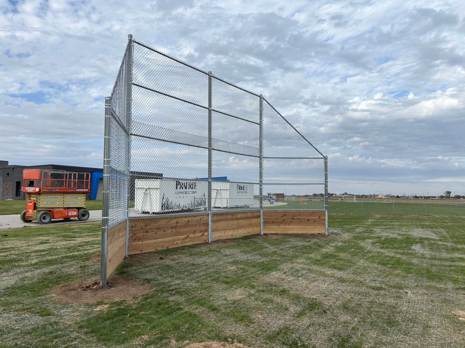 A chain-link fence with a wooden base, set on a grassy field, with construction equipment in the background.