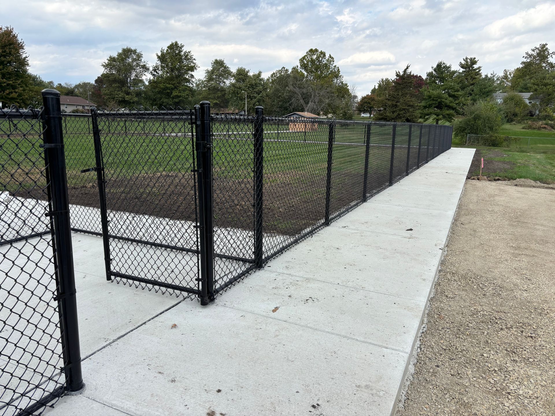 Black chain-link fence bordering a concrete walkway. Trees and grass are visible in the background under a cloudy sky.
