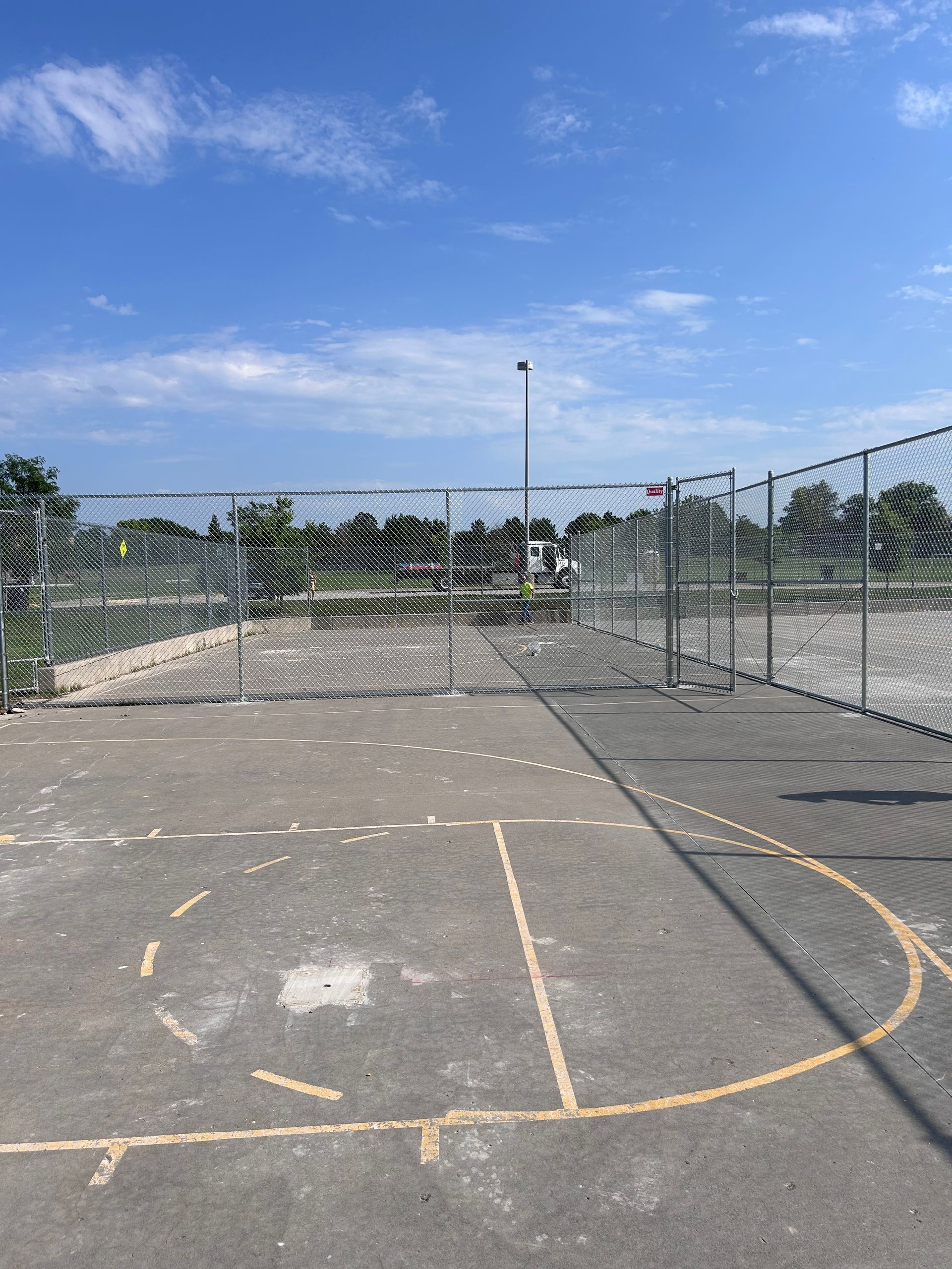 Empty outdoor basketball court behind a chain link fence under a blue sky.