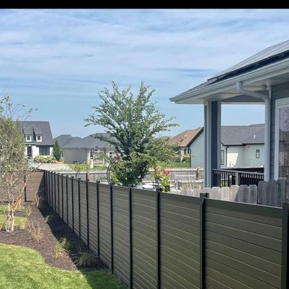 Wooden fence alongside a house and yard with a tree and other houses in the distance under a blue sky.