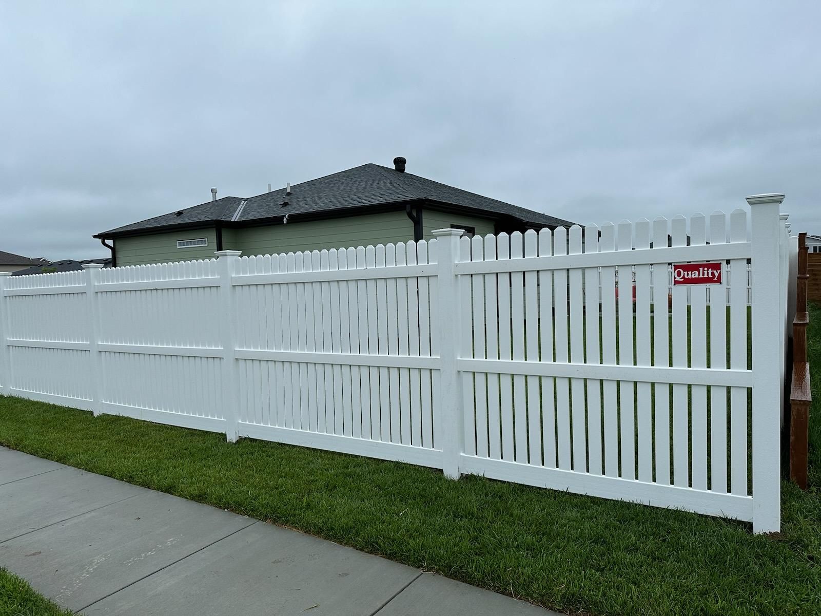 White picket fence in front of a house, with green grass and a gray sky.