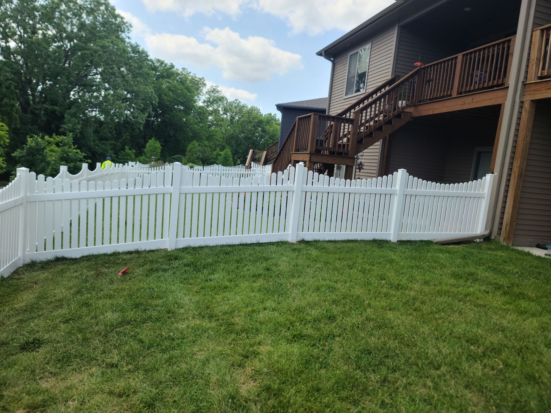 White vinyl fence in a grassy yard, partially enclosing the property of a brown house with wooden stairs.