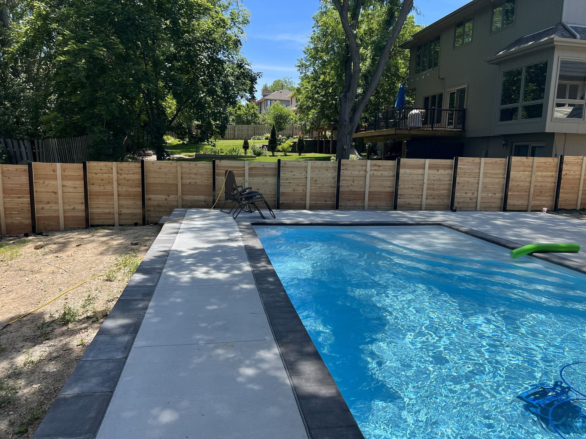A backyard pool with a concrete walkway, wooden fence, and a house in the background. Blue water.