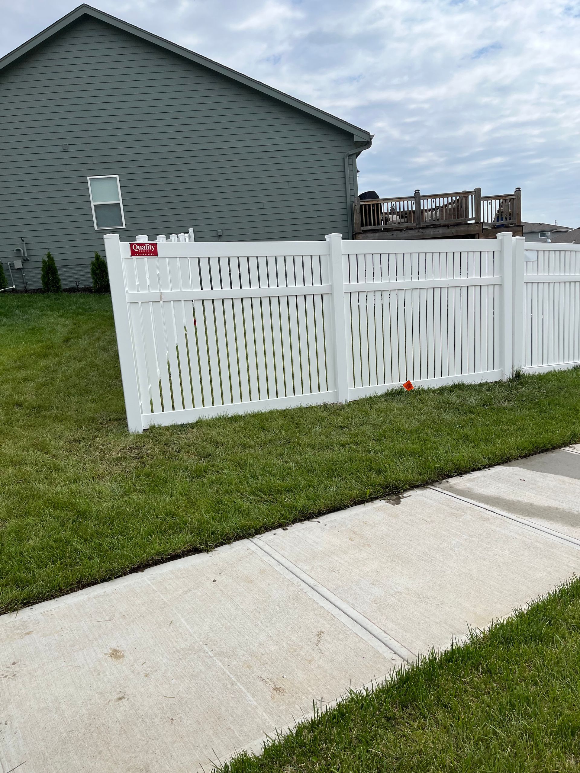 White decorative fence in a yard with green grass, next to a sidewalk and a house.