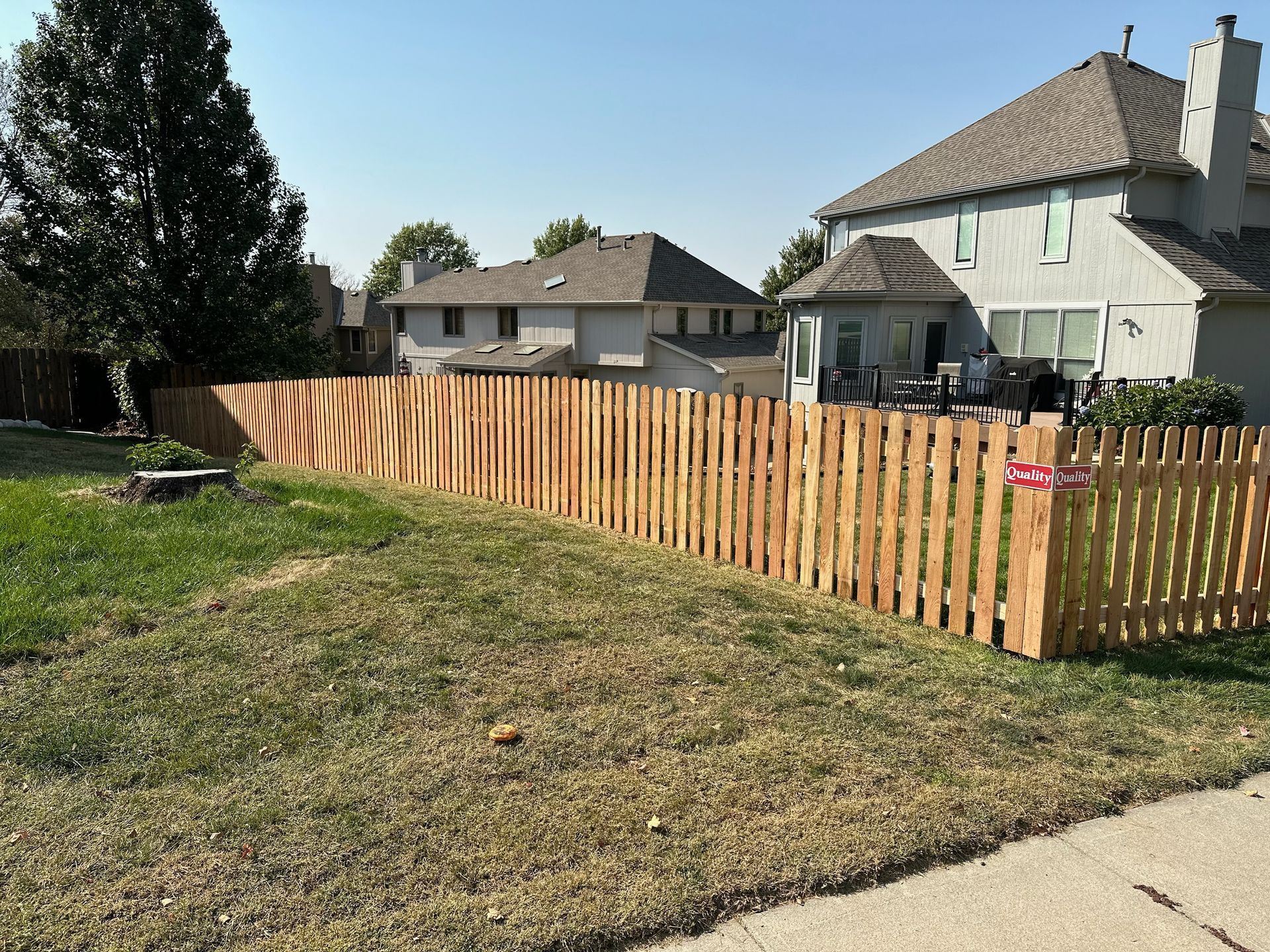 Wooden fence in a yard, separating grassy area from two-story house under a clear sky.