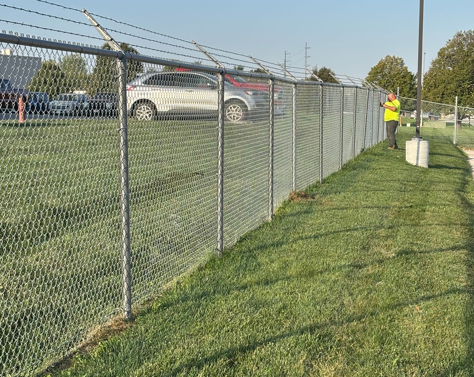 Chain-link fence with barbed wire atop, separating grassy area from a parking lot. A person in a vest stands near it.