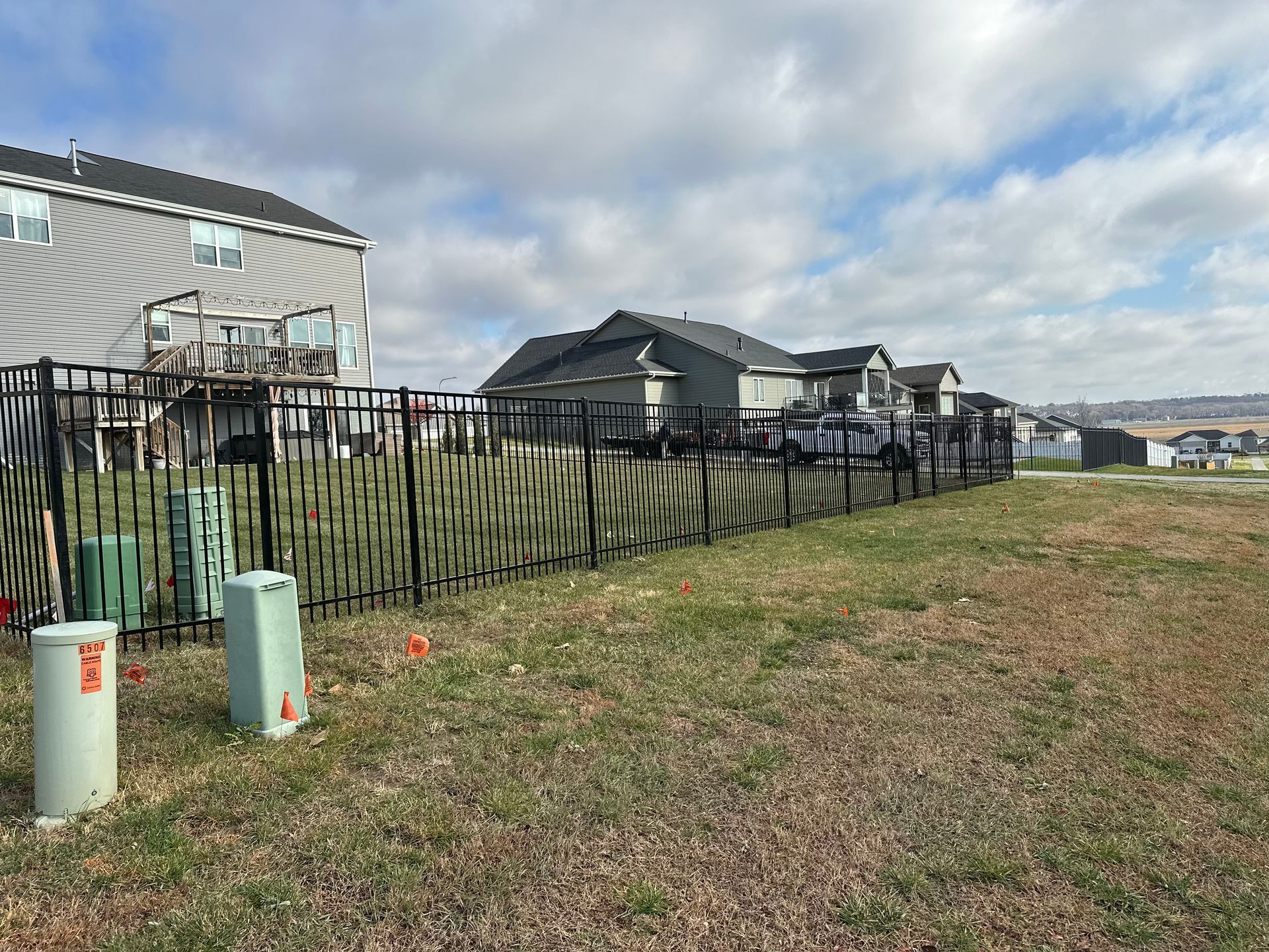 Black metal fence bordering a grassy area with houses and a cloudy sky.