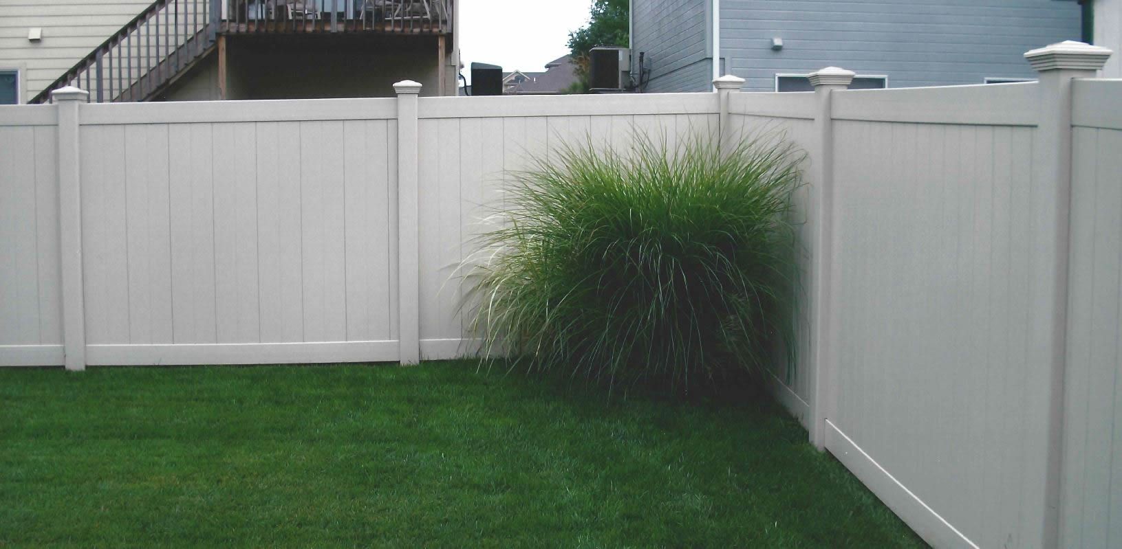 White vinyl fence encloses a green lawn. A large green plant sits next to the fence. Buildings in the background.