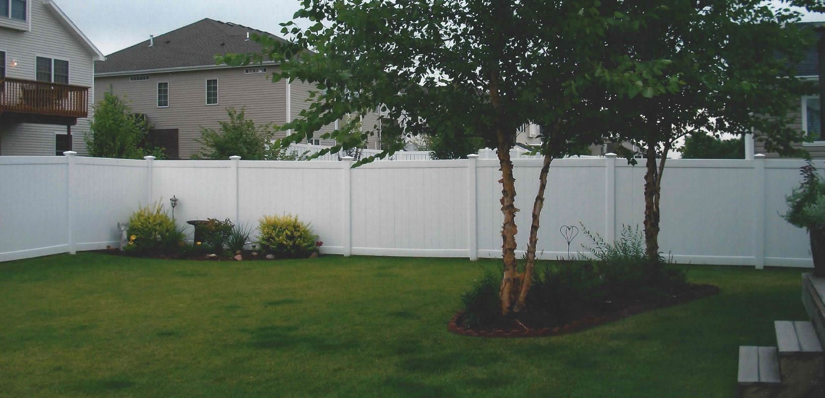 A backyard with a white fence, green grass, and trees. Two houses are visible in the background.