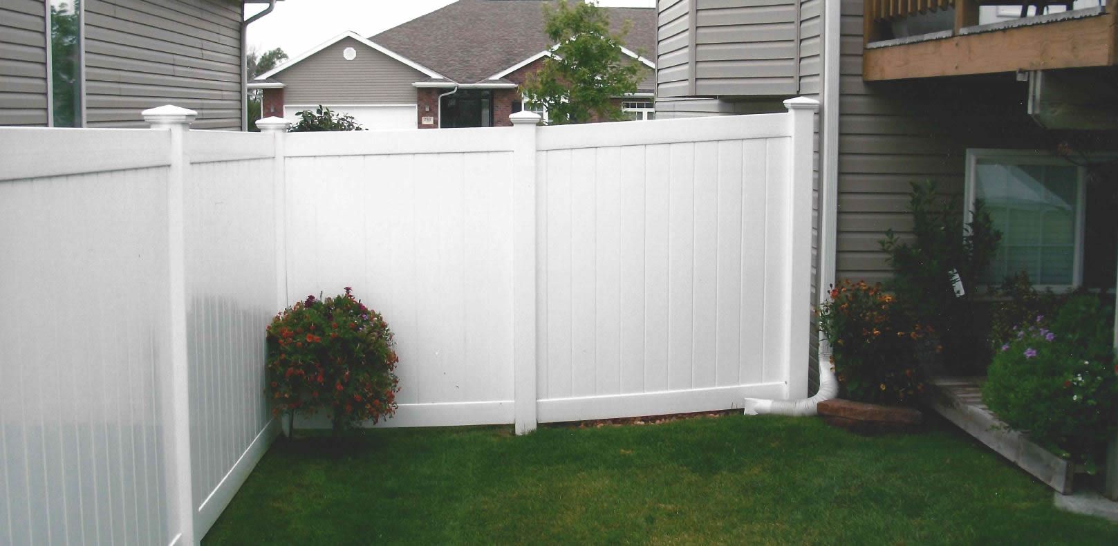White vinyl fence in a grassy backyard, adjacent to a house with siding and another building.