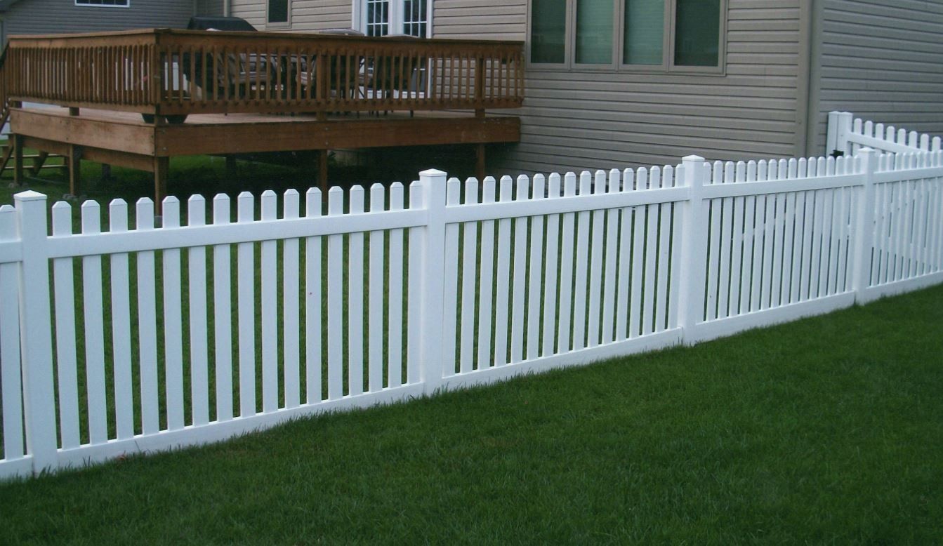 White picket fence in a green yard, adjacent to a wooden deck and light-colored house.
