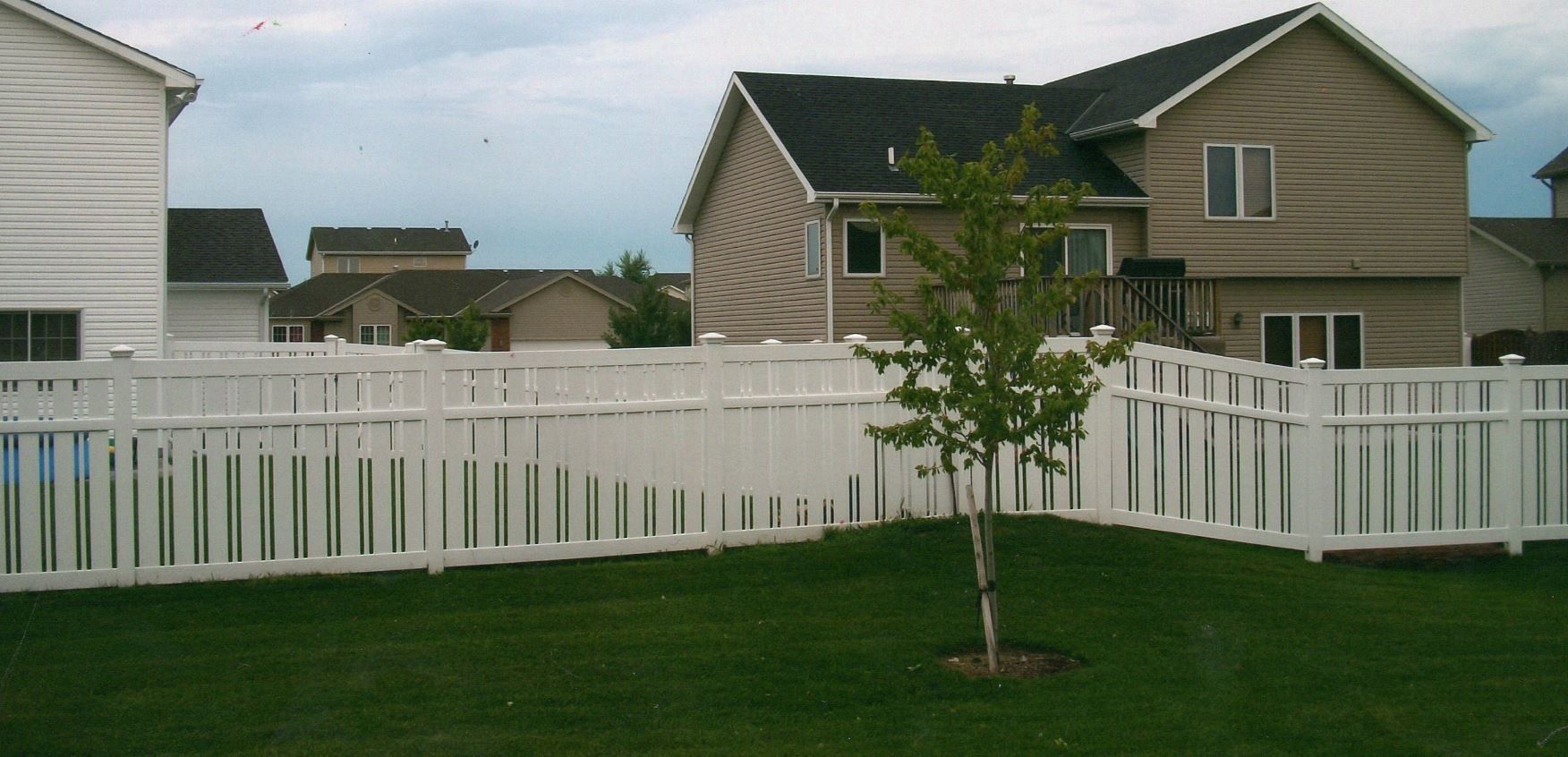 A backyard with a white fence, green grass, a small tree, and houses. Overcast sky.
