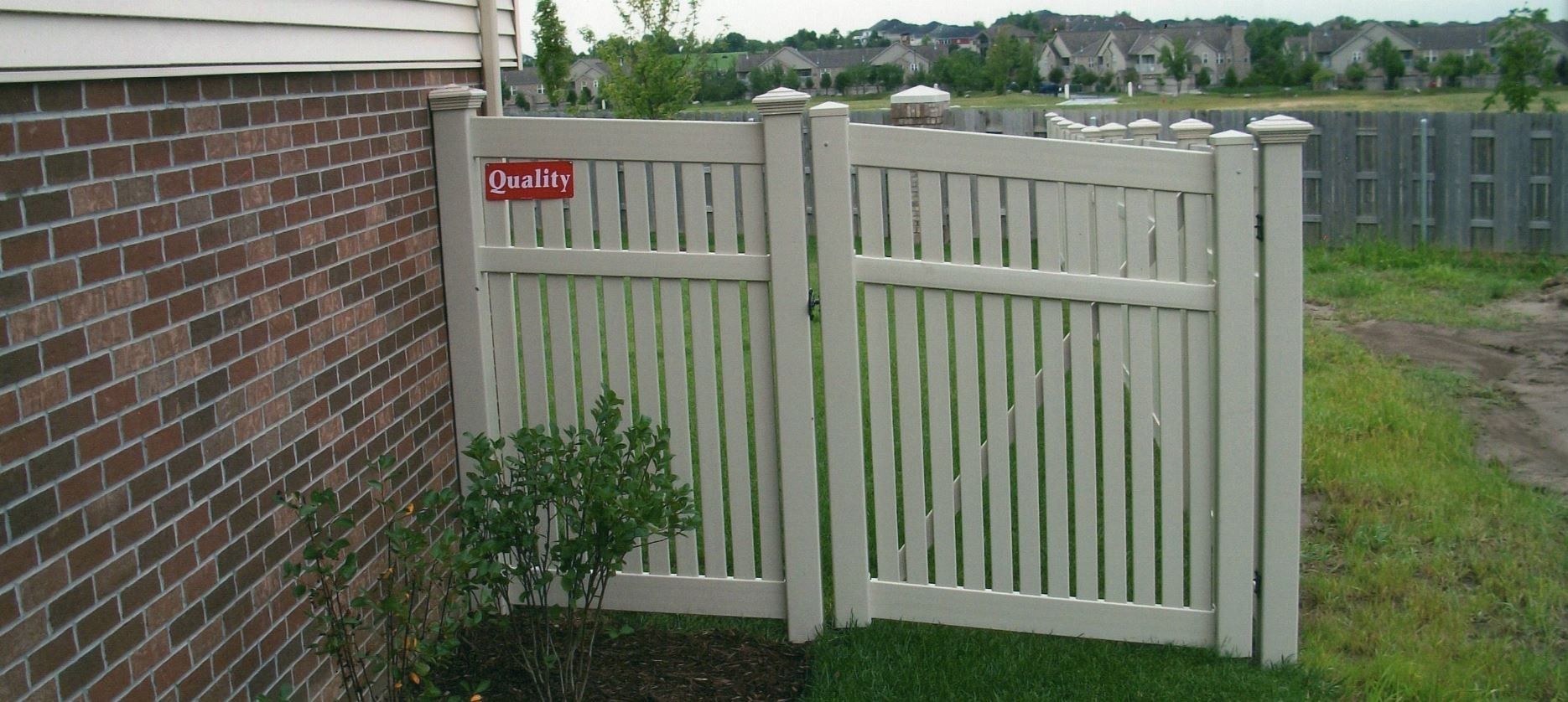Beige picket fence gate next to a brick wall. Green grass and small bush in front.