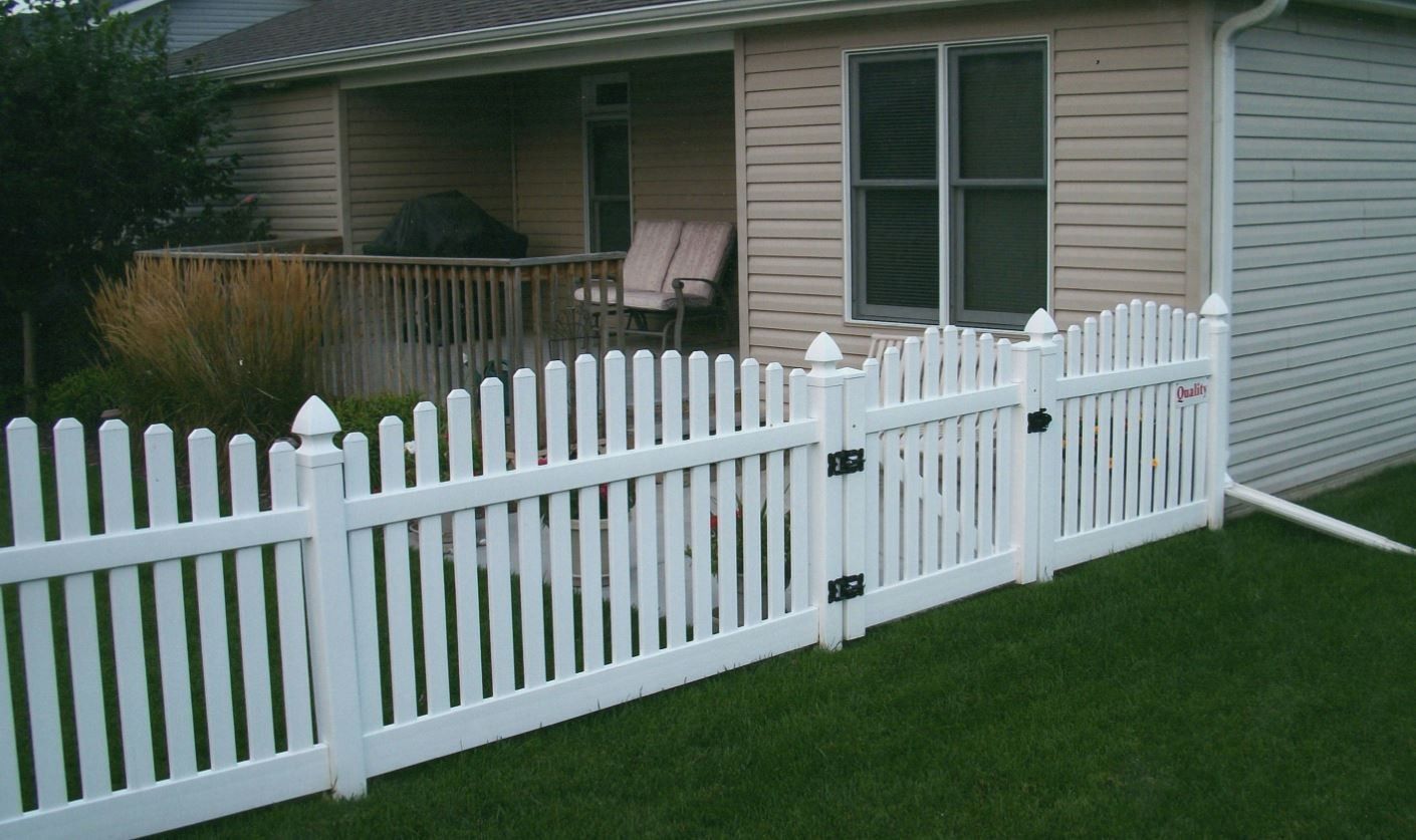 White picket fence with gate in front of a house. Green grass in foreground, siding on the house is beige.