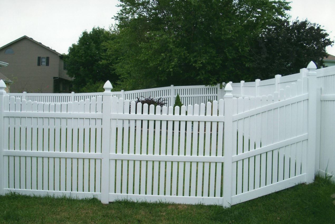 White picket fence encloses a backyard with a house in the background. Green trees and grass.