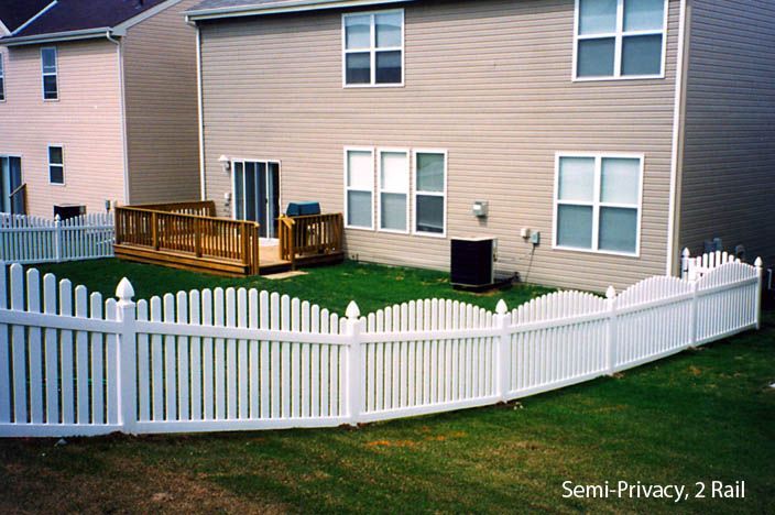 White picket fence surrounds a backyard with a wooden deck and tan house.