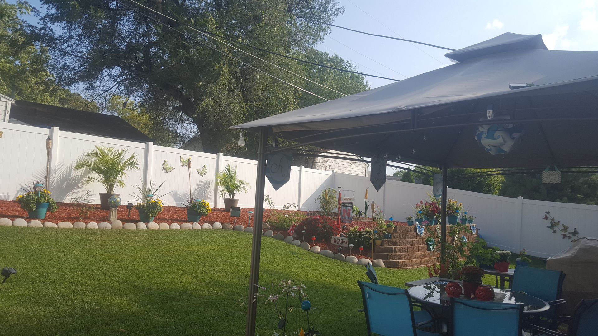 Backyard with white fence, gazebo, green lawn, flower beds, and string lights.
