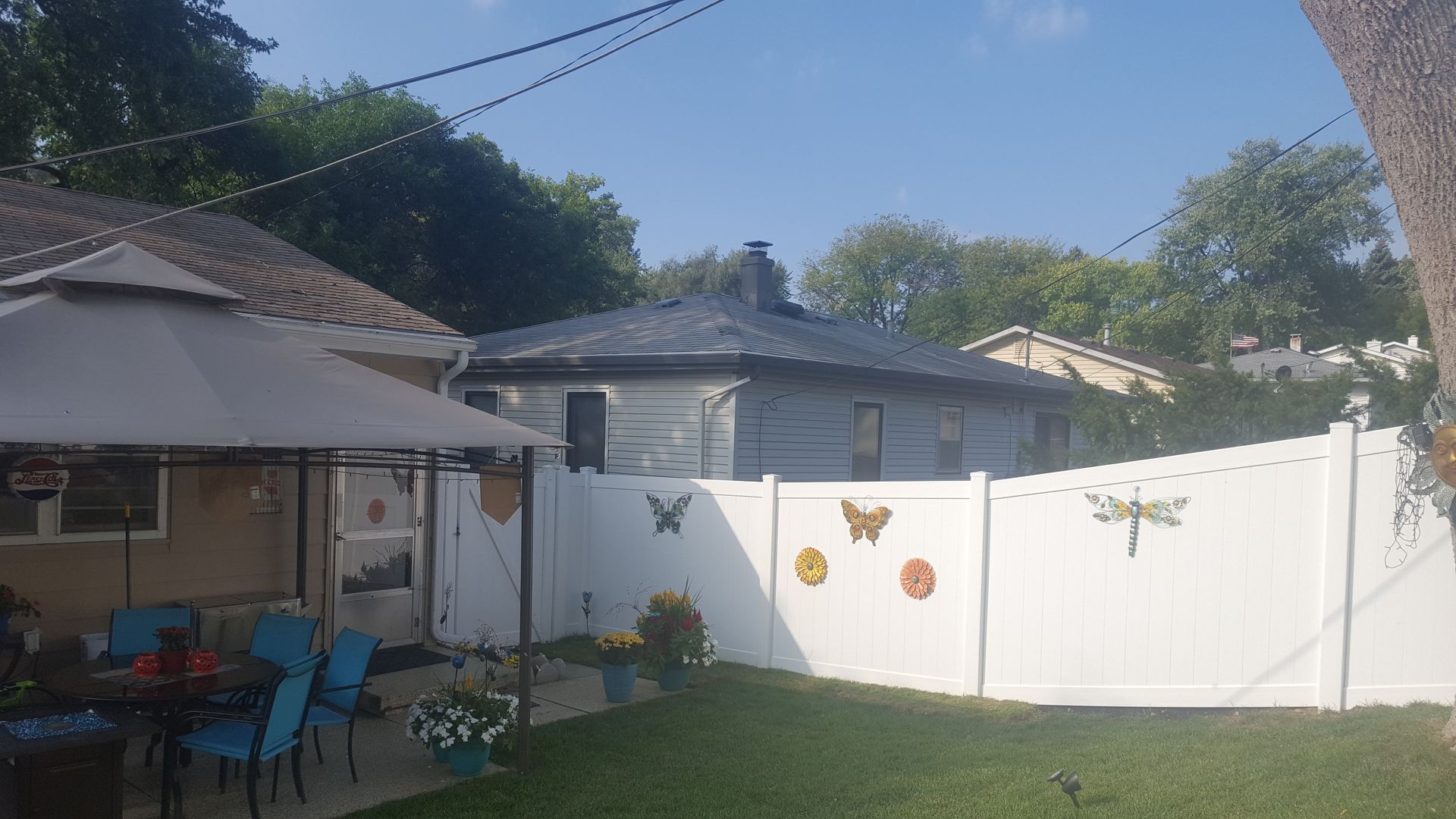 Backyard with white fence, blue chairs, patio umbrella, and decorations; trees and houses in the background.