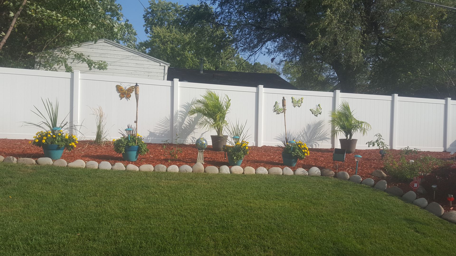 A white fence with butterfly decorations, flower pots, red mulch, and a green lawn.