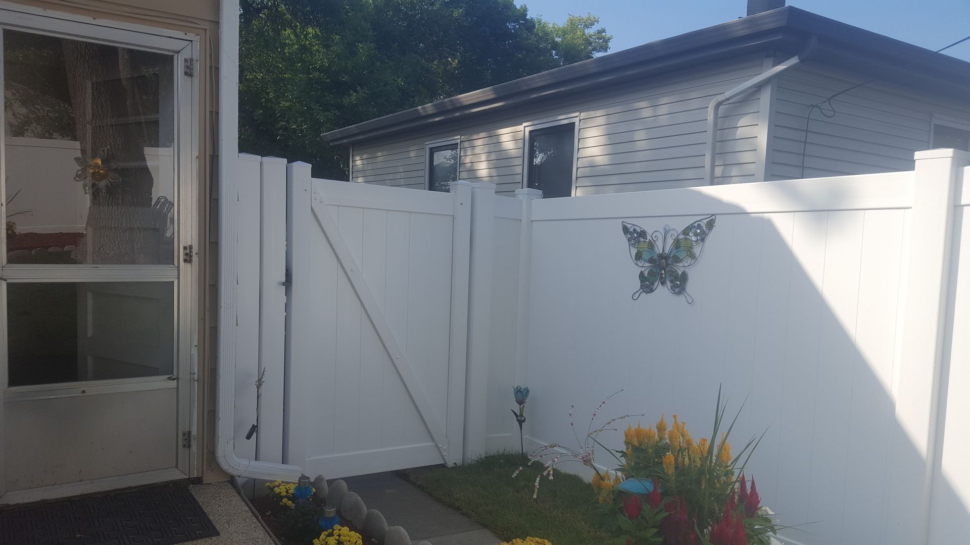 White fence with a gate, next to a doorway, and a house in the background. Butterfly decoration.
