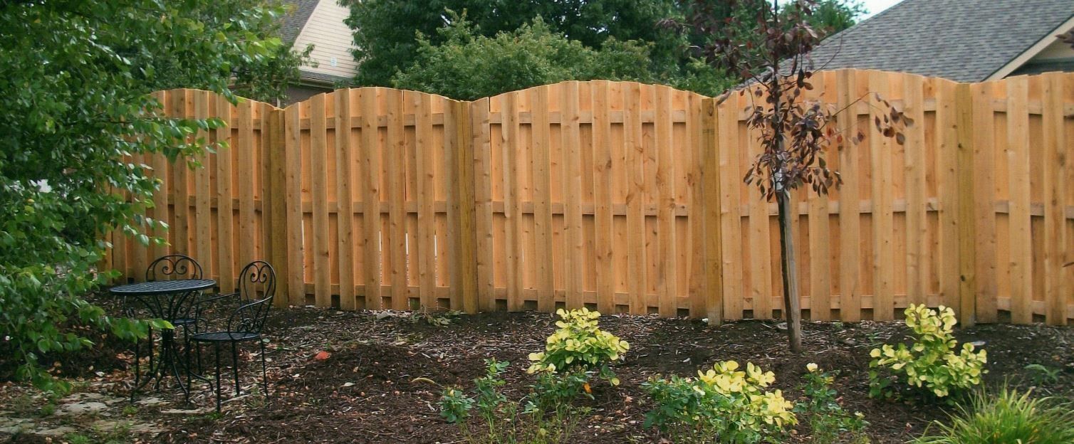 Wooden fence with arched top in a yard with trees and bushes.