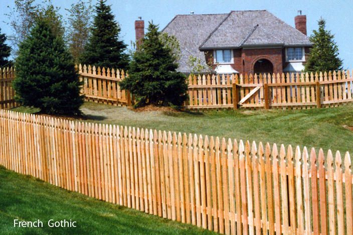 Wooden French Gothic fence surrounding a grassy hill with a house.