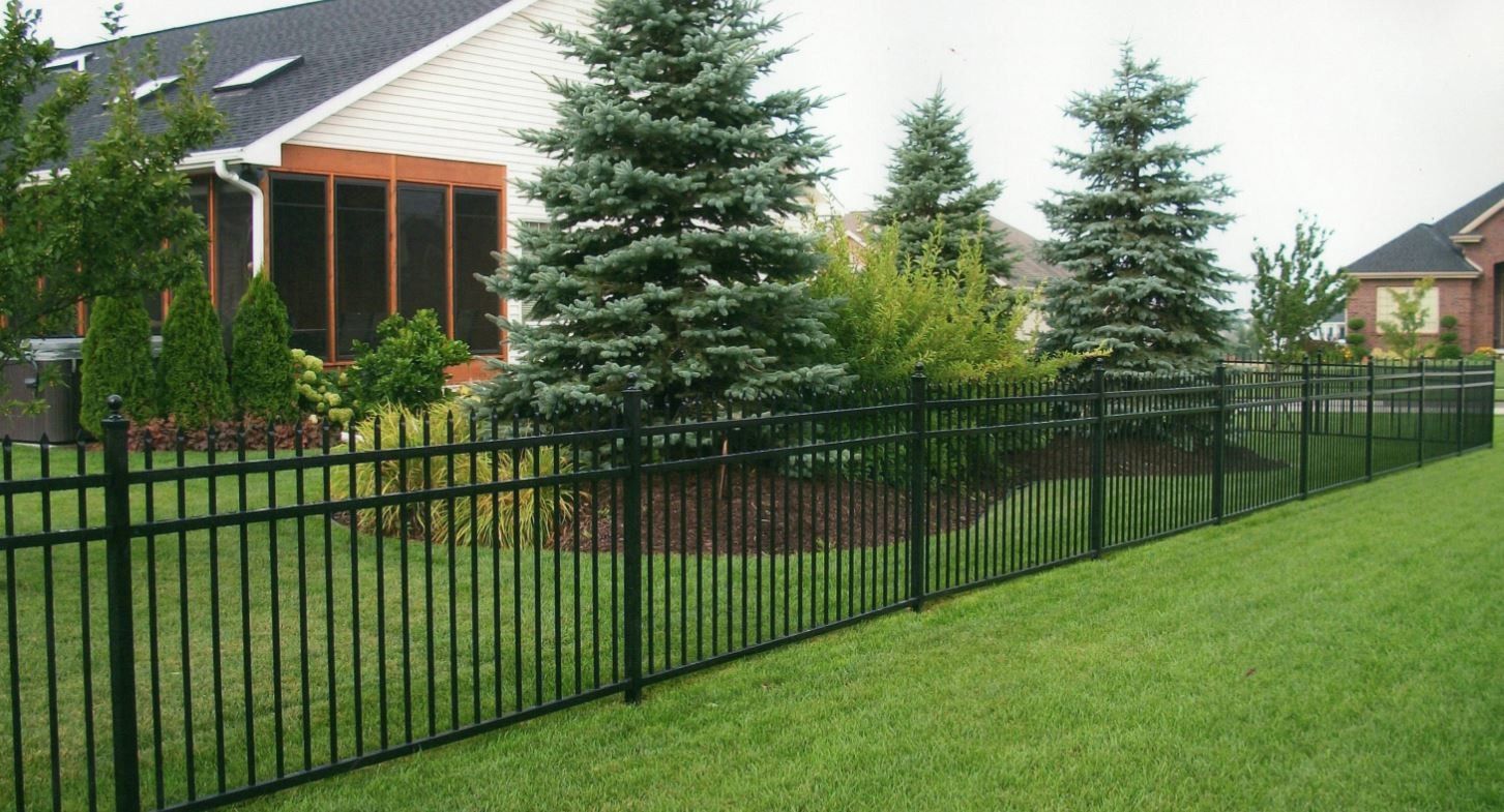 Black metal fence along a grassy yard, with evergreen trees and a house in the background.
